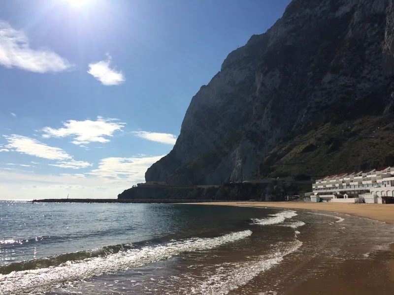 Písečná pláž, moře a strmá stěna Gibraltarské skály
