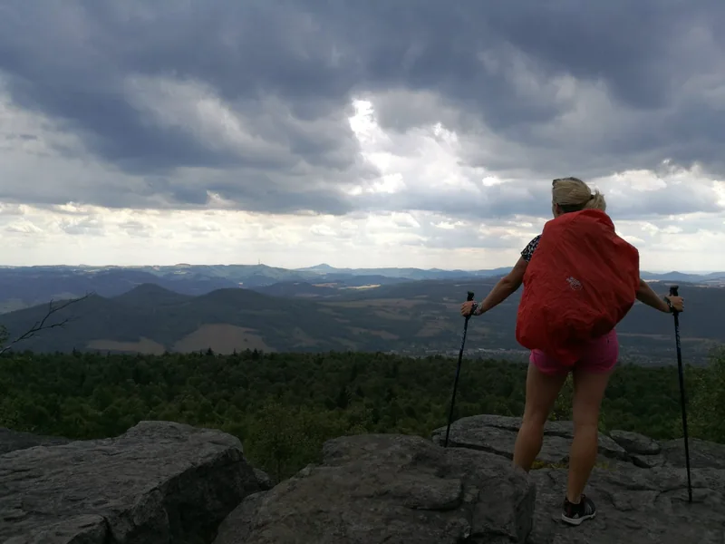 Turistka zády k fotoaparátu s červeným batohem a holemi na vyhlídce pod mraky