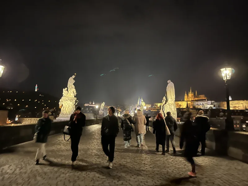 Večerní Karlův most se sochami, rozsvícenými lampami a Pražským hradem v pozadí